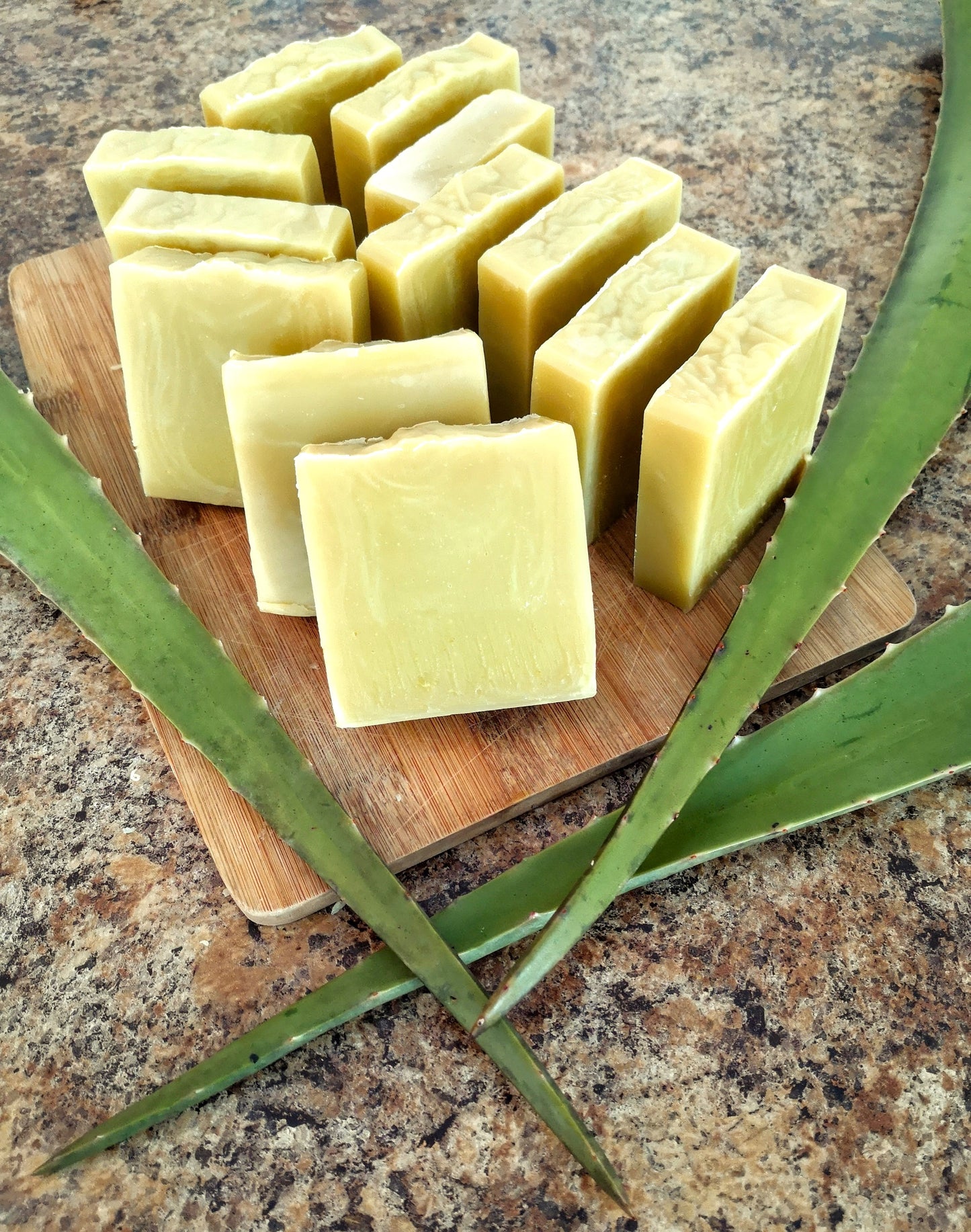 12 bars of yellow/green soap arranged on a wooden cutting board. There are three aloe leaves arranged around the soap. All on a brown and black specked counter top.