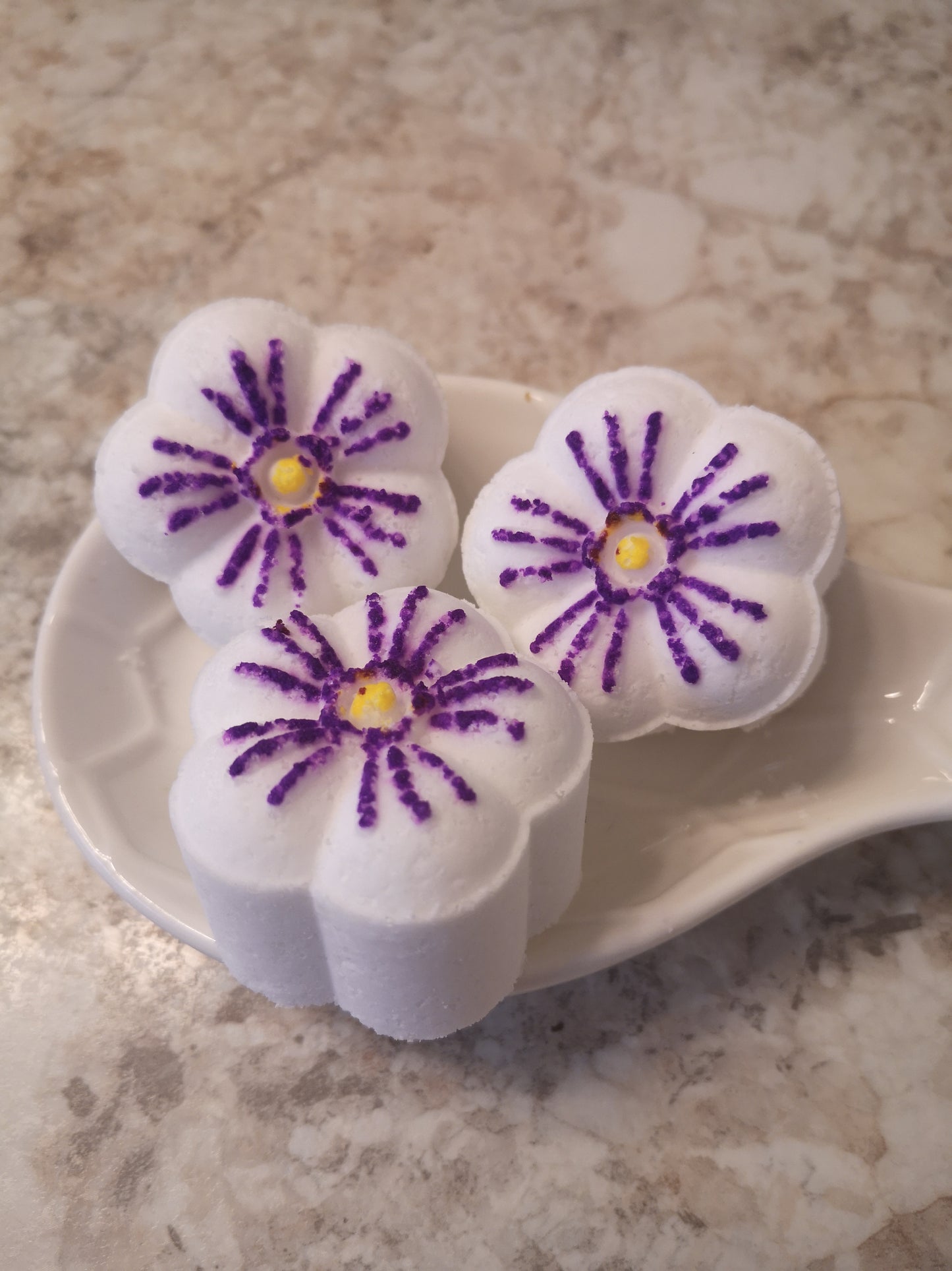 Three white flower shaped steamers with purple flower petals and yellow centres on a white ceramic spoon. Resting on a brown speckled counter top. 