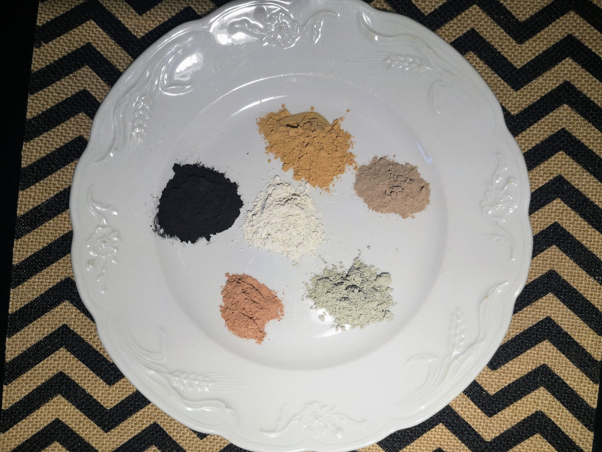 White porcelain plate on a chevron background. There are 6 small piles of clay on the plate: charcoal, caramel, rhassoul, kaolin, peach and green.