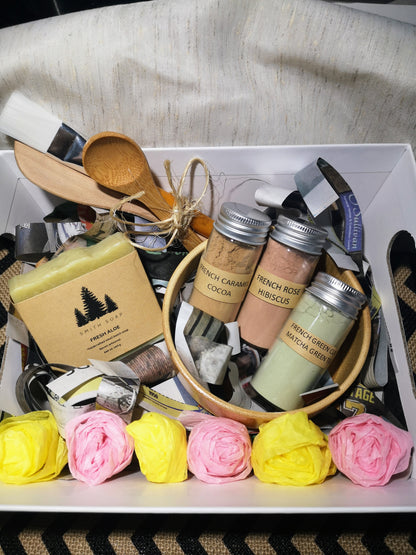 A picture of the gift basket with contents. White box containing one bar of light green soap wrapped in soap label; set of spoon, spatula and brush tied with sissal, one bamboo bowl containing three glass jars with clays in them. The basket is decorated with yellow and pink crepe paper rosettes. 
